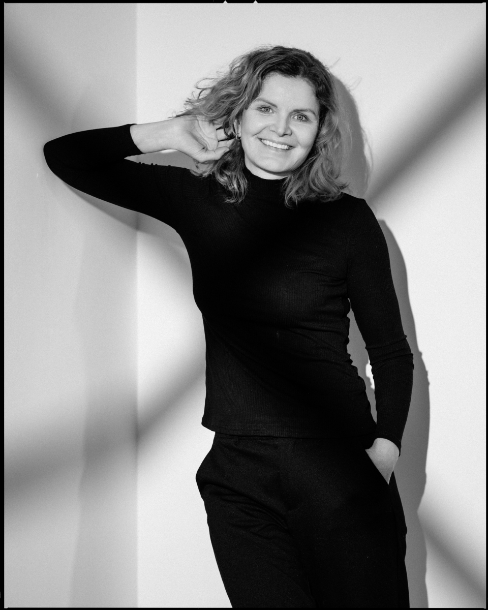 Frau mit lockigem Haar im Rollkragenpullover in Schwarz-Weiß, vor einer Wand stehend.