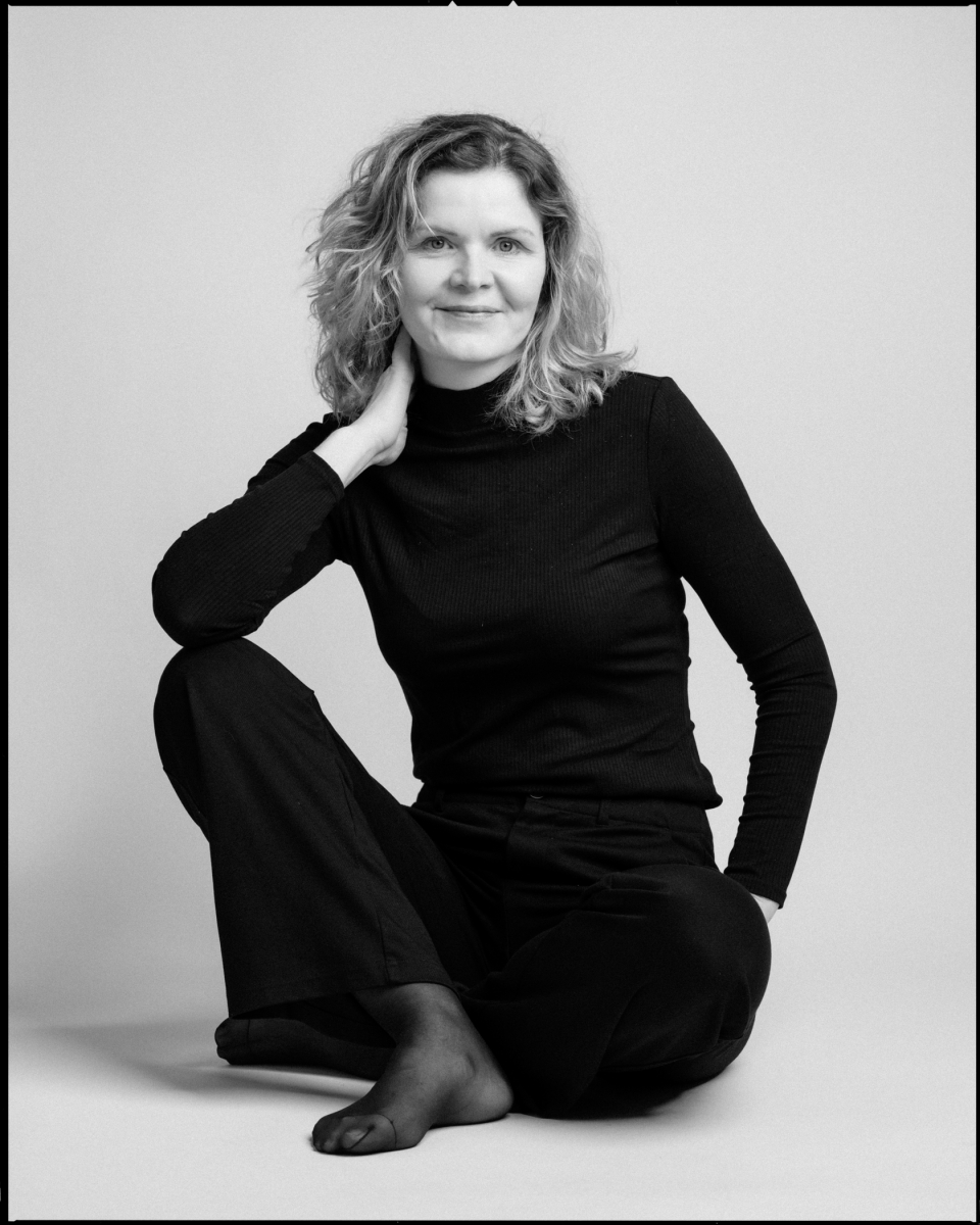 Person mit lockigem Haar in schwarzer Kleidung sitzt auf Boden und schaut in die Kamera.