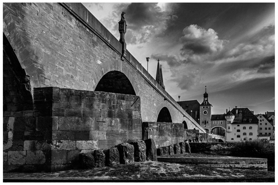 Historische steinerne Brücke mit Bögen, Fluss, traditionelle Gebäude, Uhrturm, wolkiger Himmel.