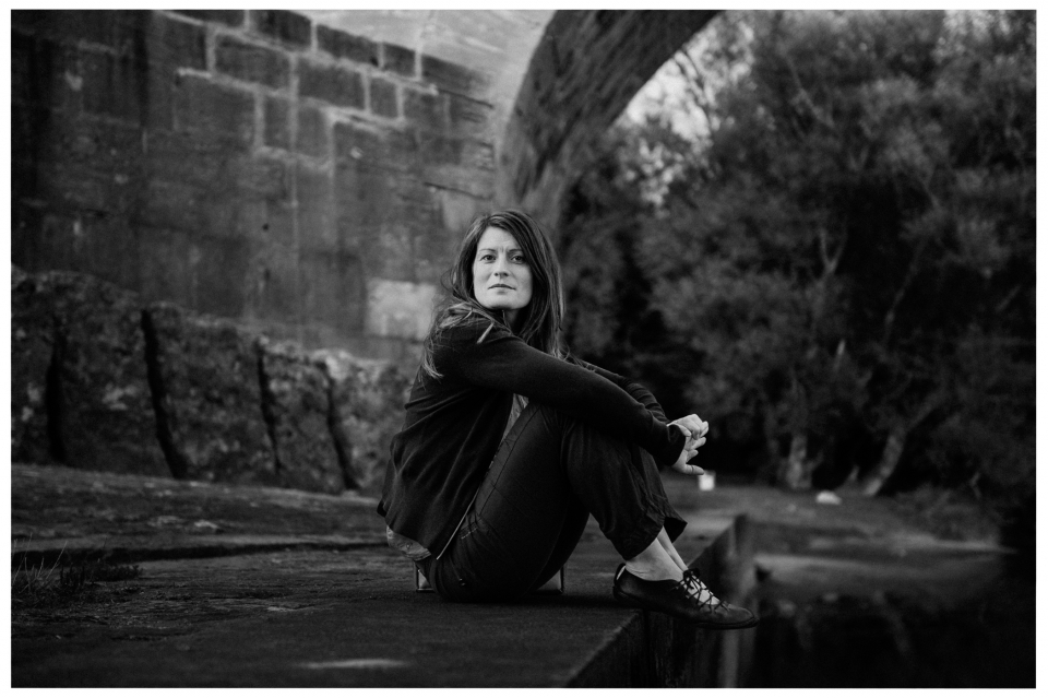 Frau sitzt nachdenklich unter Brücke, Bäume im Hintergrund, schwarz-weiß, ruhige Atmosphäre