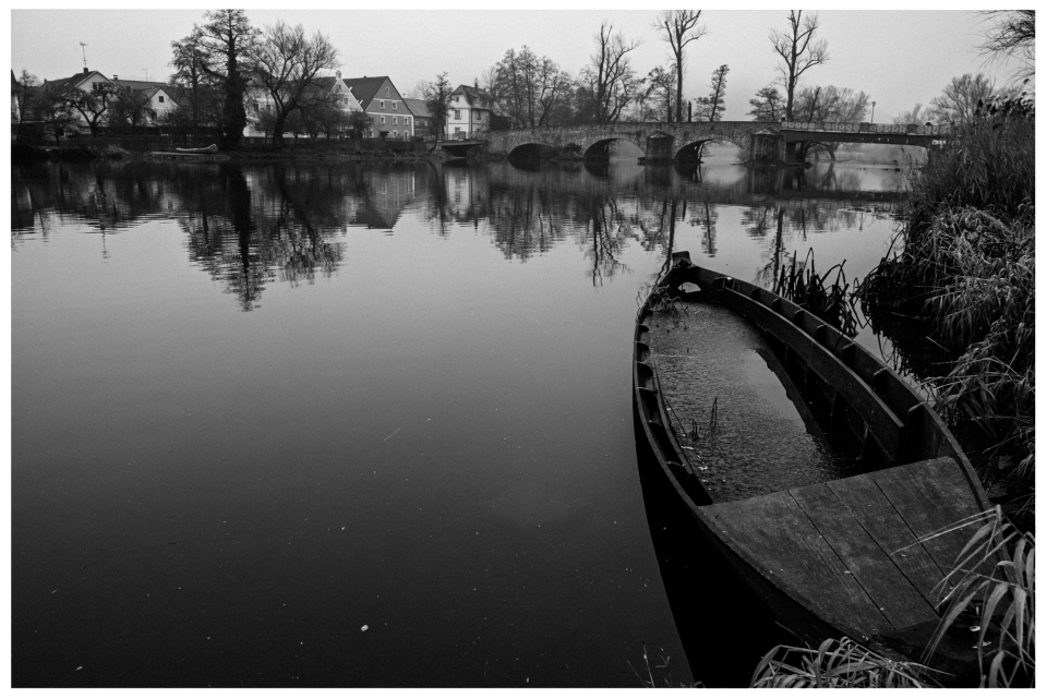 Ruhiger Fluss mit alter Bootsanlegestelle, steinerne Brücke und Häuser im Hintergrund.