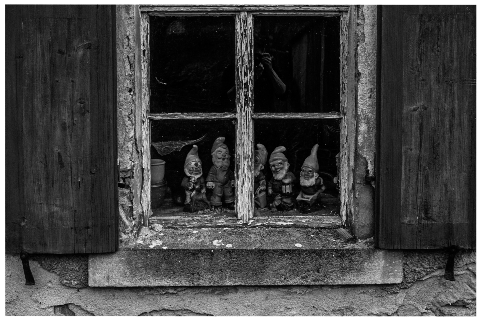 Verwitterte Fensterbank mit Gartenzwergfiguren hinter abgenutzten Holzläden und gealtertem Glas.