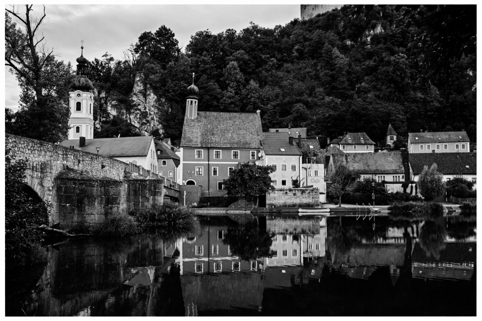 Dorf am Fluss mit Kirche, Zwiebelturm und bewaldeten Hügeln, Gebäude spiegeln im Wasser.