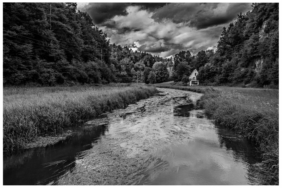 Fluss durch bewaldete Landschaft mit Haus am Ufer und bewölktem Himmel.