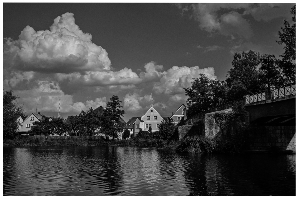 Traditionelle Häuser und Brücke am Flussufer unter bewölktem Himmel, flankiert von Bäumen.