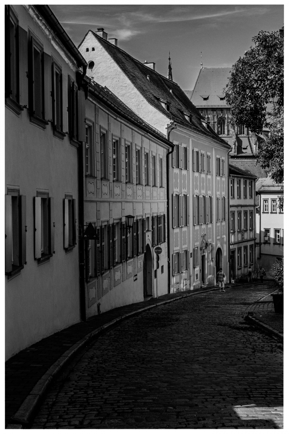Historische gepflasterte Straße mit Gebäuden, shuttered Fenstern, Kirche im Hintergrund, Menschen.