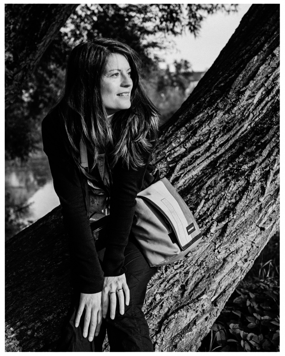 Person sitzt lächelnd auf Baumstamm in der Natur; Schwarz-Weiß-Fotografie mit Umhängetasche.