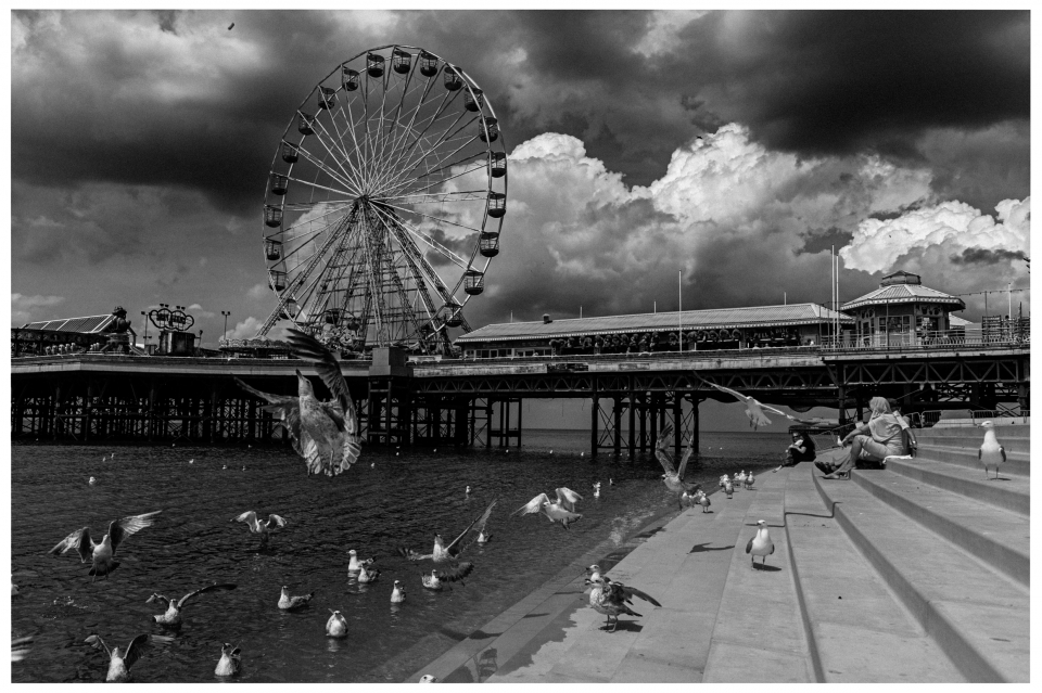 Riesenrad auf einem Pier, umgeben von Möwen und dunklen Wolken, Menschen auf Stufen sitzend.
