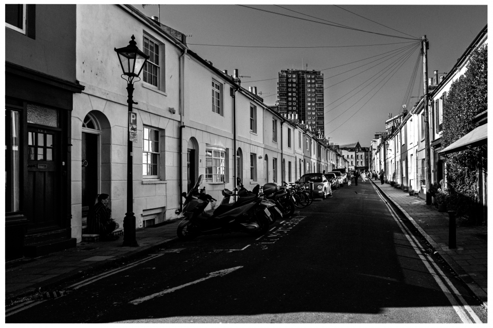 Schwarz-weiß Foto einer städtischen Straße mit Motorrädern, Autos, Reihenhäusern und Hochhaus.