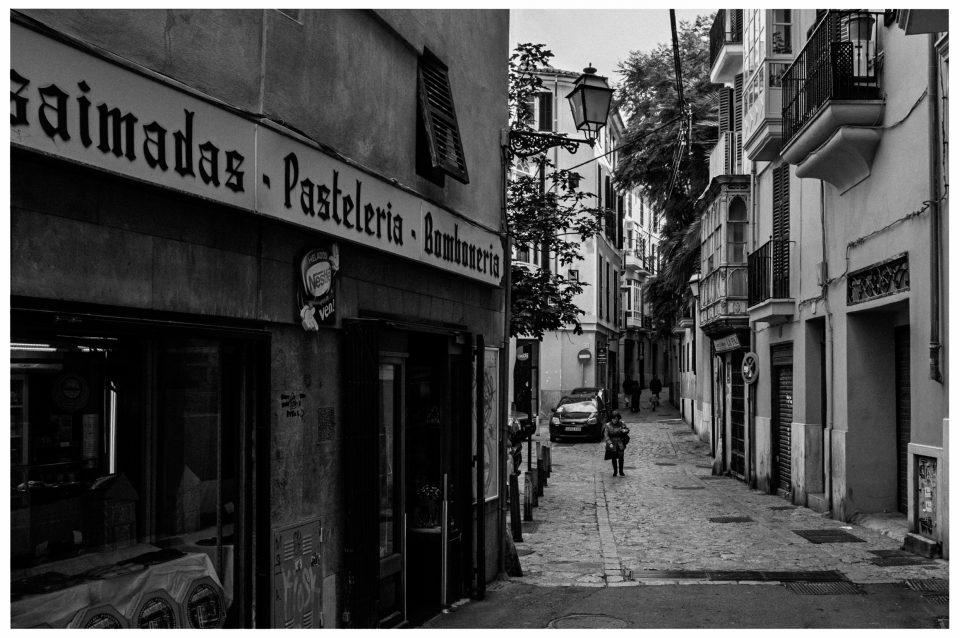 Schwarzweißfoto einer kopfsteingepflasterten Straße mit Geschäften und Balkonen in einer Stadt.
