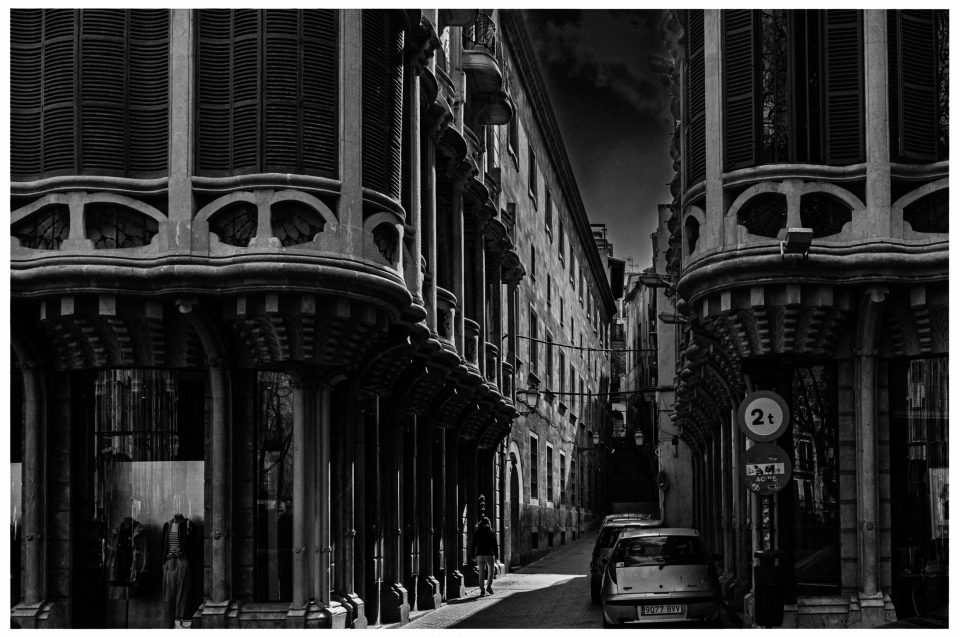 Enge gepflasterte Straße, markante Gebäude, geparkte Autos, wenige Fußgänger, Schwarz-Weiß.