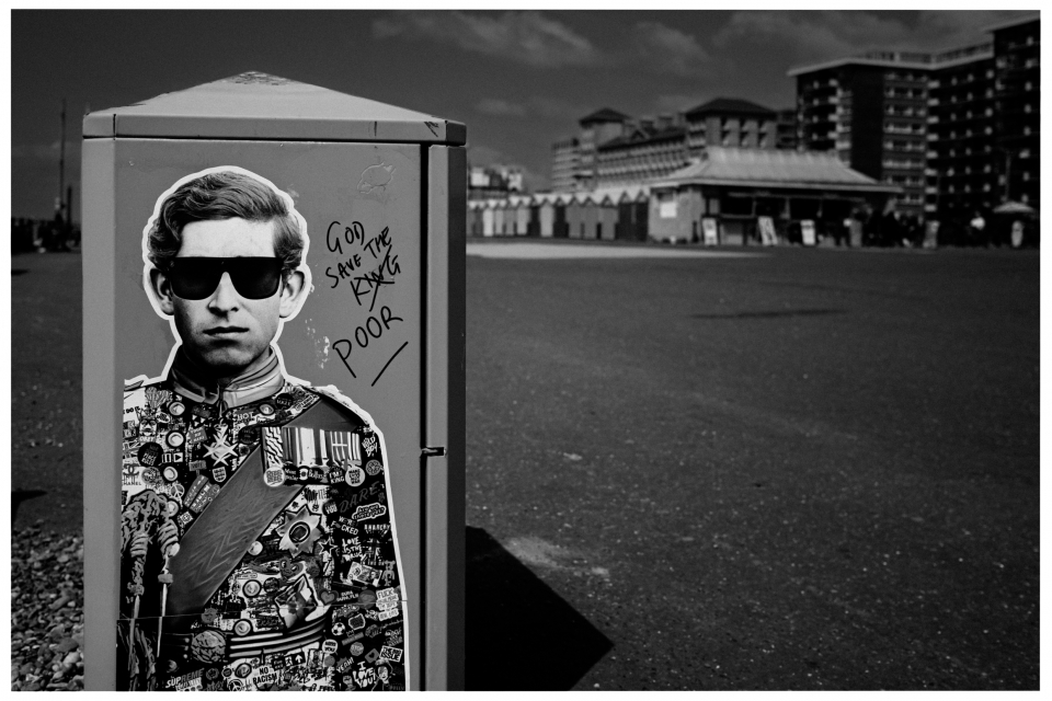 Stromkasten mit Graffiti einer uniformierten Figur, handschriftliche Worte, Gebäude im Hintergrund.