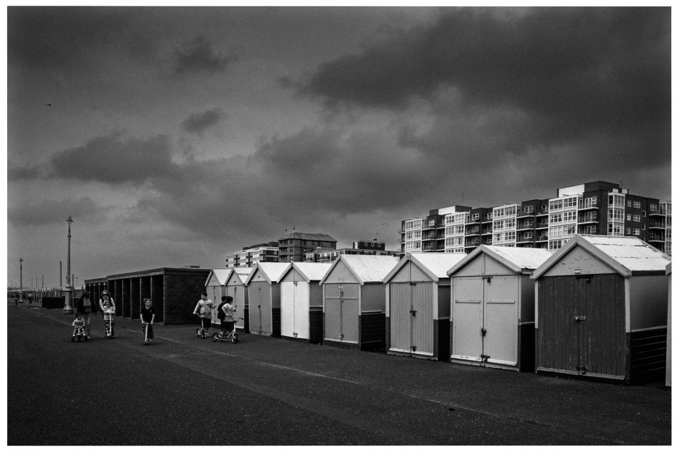 Reihe von Strandhütten an einer Promenade, Mehrfamilienhäuser im Hintergrund, wolkiger Himmel.