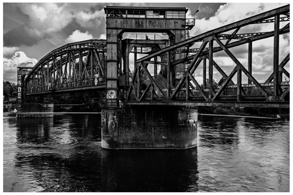 Stählerne Brücke mit komplexen Metallstreben überquert Fluss, Himmel voller Wolken.