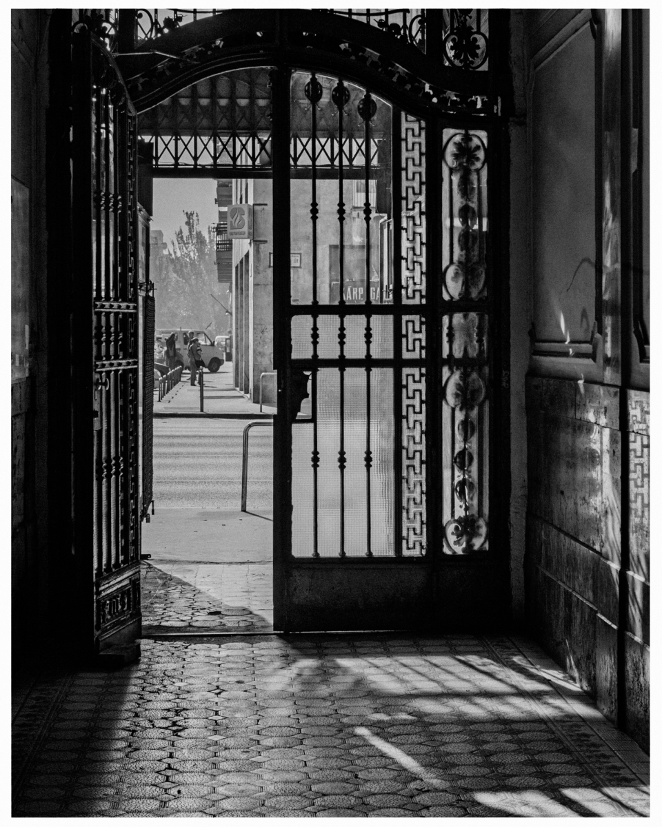Eisengittertor öffnet sich zu sonnendurchfluteter Straße mit Schattenmustern und Gebäuden.