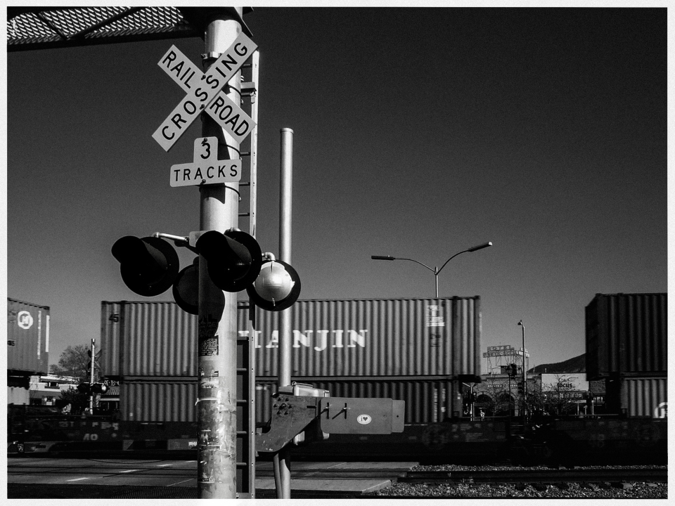 Bahnübergang mit Schild "3 Gleise", Güterwaggons im Hintergrund.
