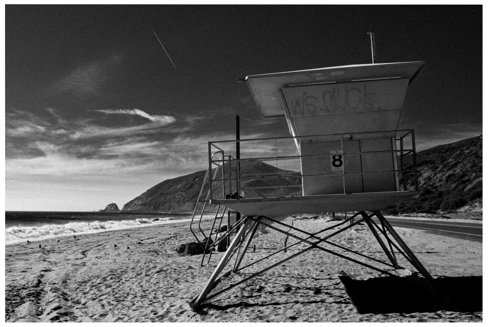 Rettungsturm Nr. 8 an einem sandigen Strand vor Hügeln und klarem Himmel mit Wolken.