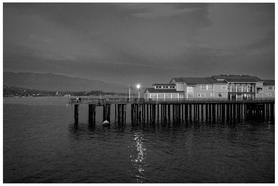 Steg über Wasser mit beleuchtetem Gebäude, Schwarz-Weiß, Berge im Hintergrund.