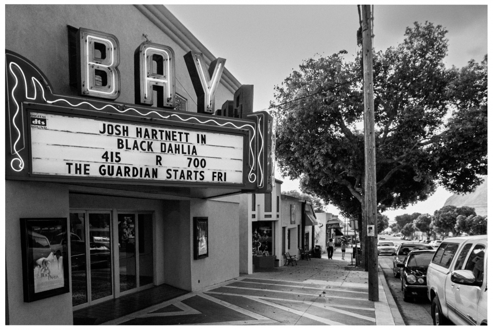 Kino mit Neonschild über aktuelle Filme, parkenden Autos und Personen auf Gehweg, schwarz-weiß.
