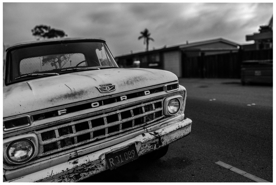 Älterer Ford-Lkw mit Gebrauchsspuren am Straßenrand, Gebäude und Baum im Hintergrund.