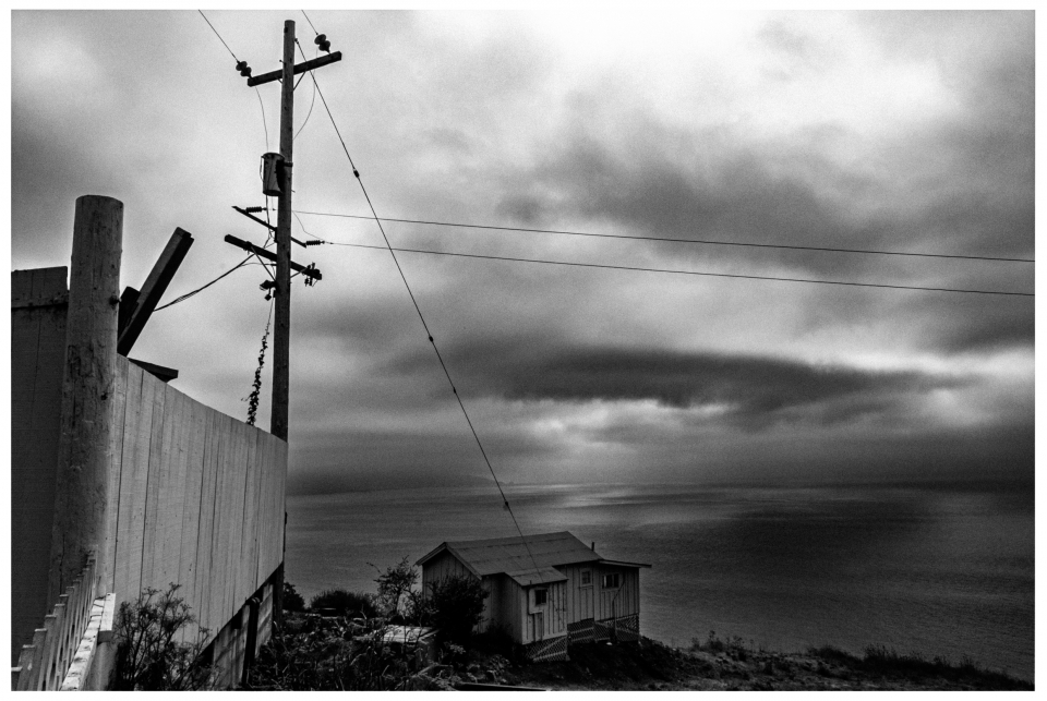 Holzhaus am Ufer mit Telefonmast, bewölkter Himmel im Schwarz-Weiß-Foto.