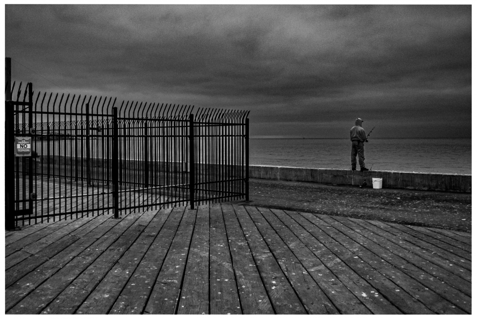 Angler steht auf Uferpromenade am Meer, Holzboden und Metallzaun mit „No Trespassing“-Schild.