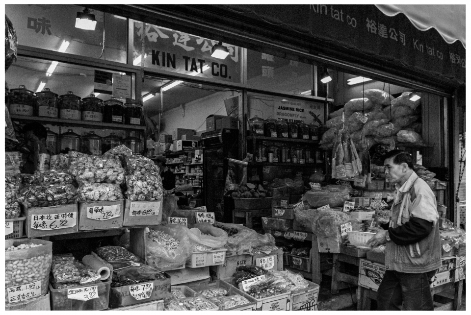 Mann vor asiatischem Lebensmittelgeschäft „Kin Tat Co.“ mit Waren, Preisschilder zweisprachig.