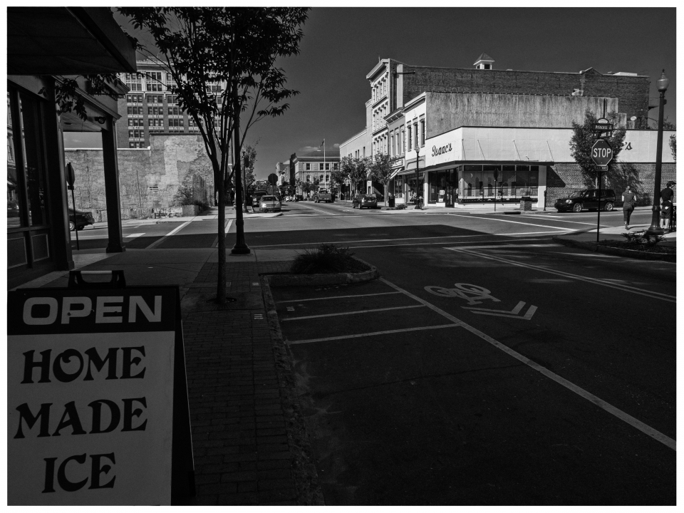 Städtische Straße mit Geschäften vor einem Backsteingebäude, Schild "HOME MADE ICE" im Vordergrund.