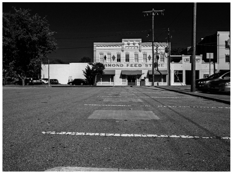 Schwarz-Weiß-Foto eines leeren Parkplatzes vor einem Gebäude mit „Feed Store“-Beschriftung.