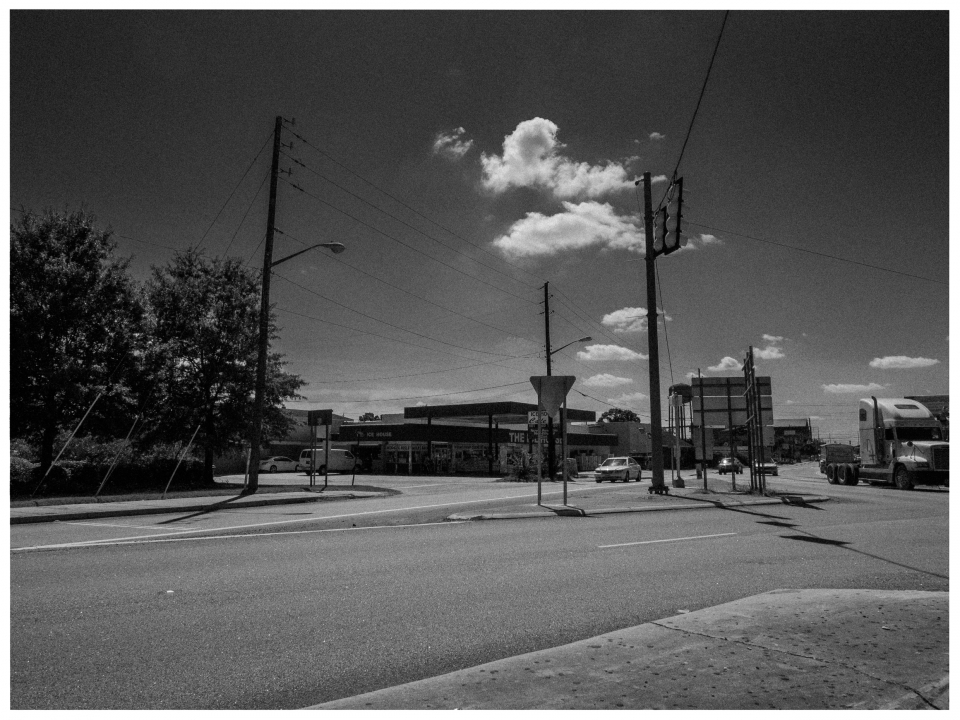 Schwarz-weiße Kreuzung mit Fahrzeugen, Bäumen, Gebäuden und bewölktem Himmel im Hintergrund.