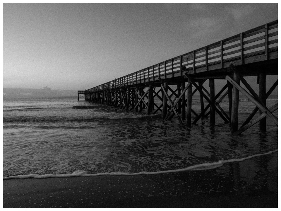 Ein hölzerner Pier ragt ins Meer, Wellen schlagen sanft gegen den Strand, gedämpftes Licht.