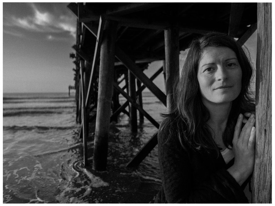 Frau unter Pier am Strand, Schwarz-Weiß, Meer und Himmel im Hintergrund sichtbar.