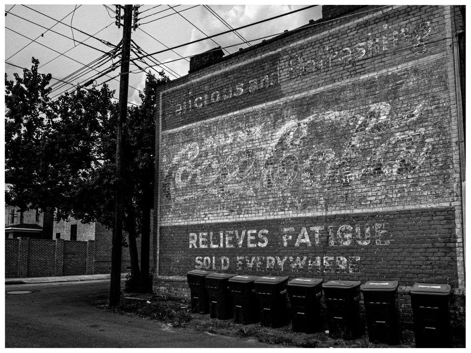 Verblasstes Getränkeschild auf Ziegelwand mit Mülltonnen darunter in städtischer Umgebung.