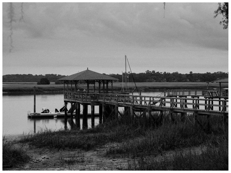 Holzsteg zu Pavillon über Wasser mit Graslandschaft und Bäumen im Hintergrund, monochrom.