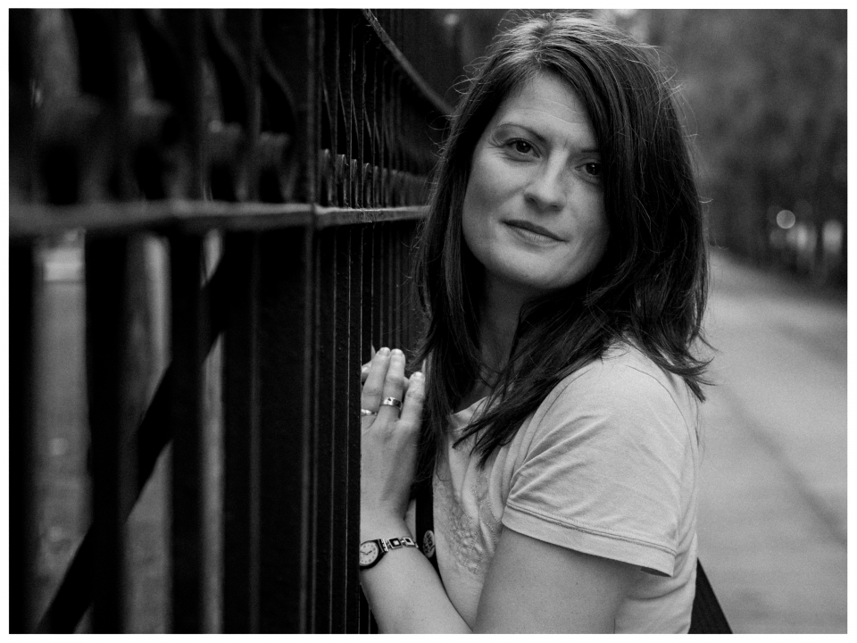 Frau lehnt an Gitter, trägt T-Shirt und Uhr, schaut mit neutralem Ausdruck in die Kamera.