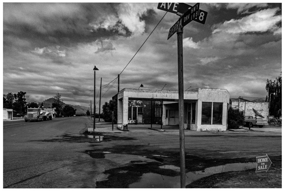 Verlassene Straßenecke, leerstehendes Gebäude, Pfützen, Lastwagen, bewölkter Himmel, Verkaufsschild.