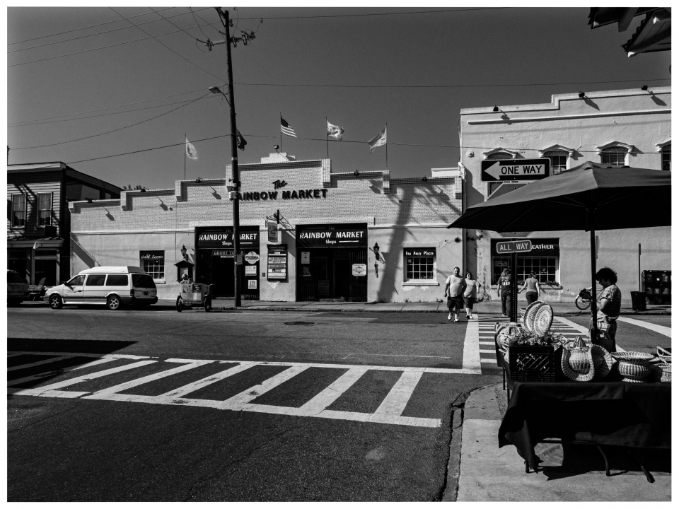 Schwarz-weiß Straßenszene mit "The Rainbow Market", Gehenden, Auto, Korbstand und "One Way"-Schild.