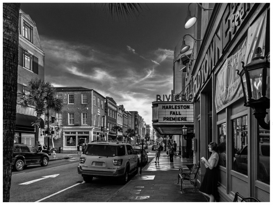Schwarz-weiße Straßenszene mit historischen Gebäuden, Kino, Autos und Menschen auf Gehweg.