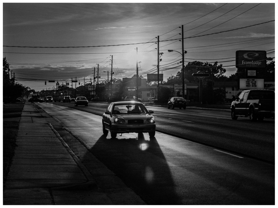 Belebte Straße bei Sonnenuntergang, Autos und Stromleitungen, Gebäude und Schilder im Hintergrund.
