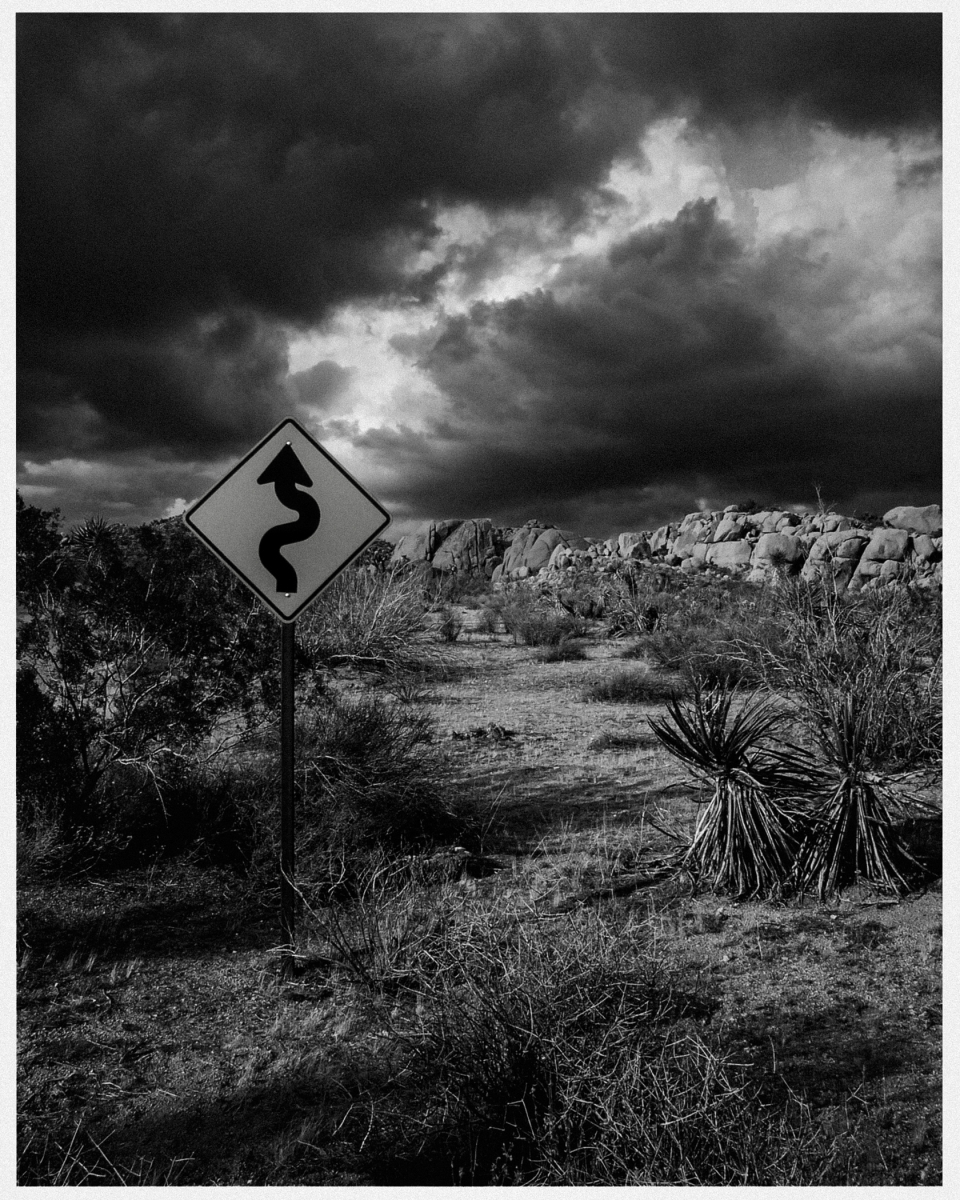 Kurvenreiches Straßenschild in trockener, steiniger Landschaft unter bewölktem Himmel.