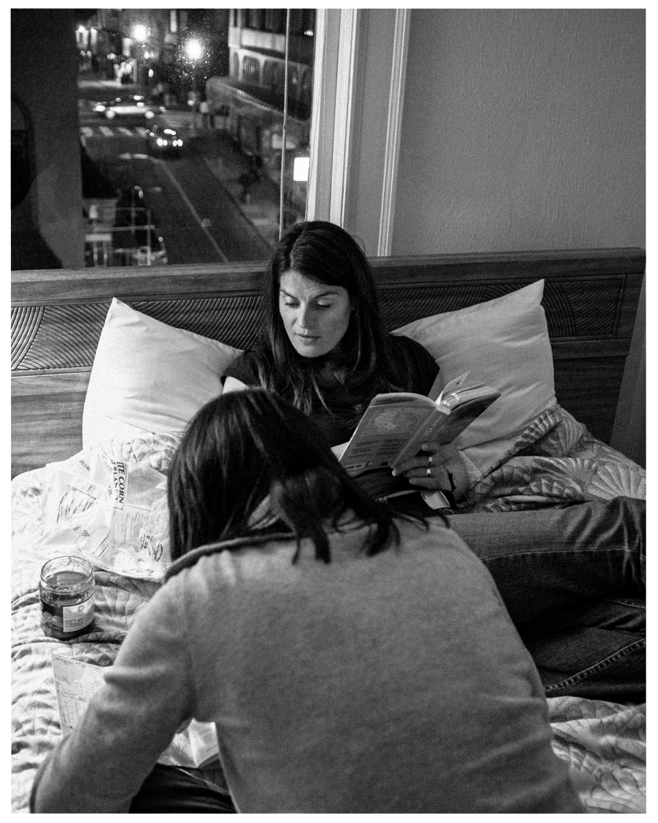 Zwei Frauen lesen auf einem Bett in einem Schlafzimmer mit Blick auf eine nächtliche Straßenszene.