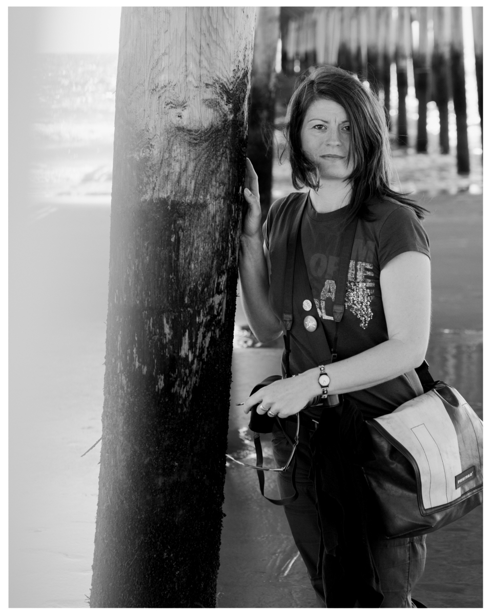 Frau am Strand neben Holzbalken, mit Tasche, schwarz-weiß, Stegpfeiler im Hintergrund.