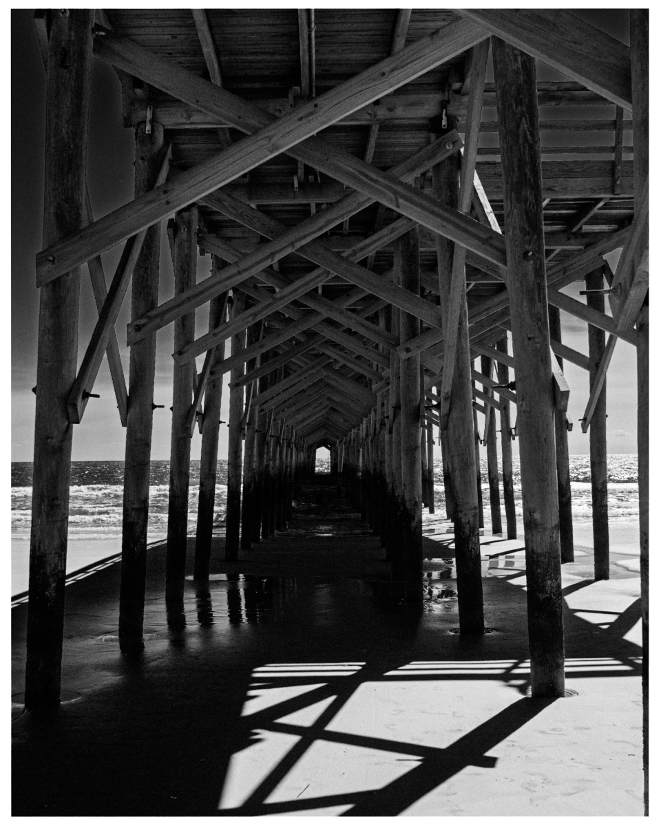 Holzbrücke über Strand mit Schatten der Balken im Sand, Meer im Hintergrund.