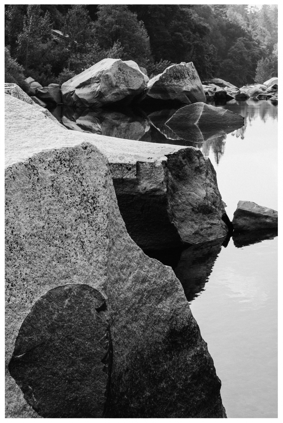 Ruhige Flusslandschaft mit spiegelnden Felsbrocken am Ufer und dichtem Wald im Hintergrund.