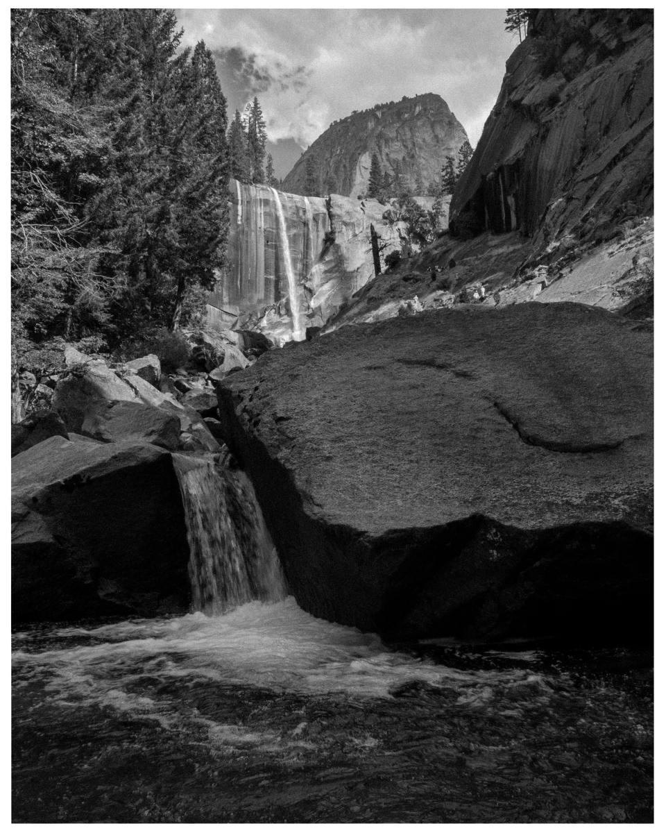 Wasserfall fließt über Felskante in einem bewaldeten Gebiet, Schwarz-Weiß-Landschaft.