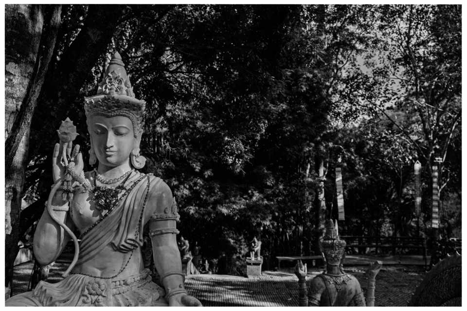 Steinfigur einer sitzenden Gottheit in traditioneller Kleidung, umgeben von Bäumen im Freien.