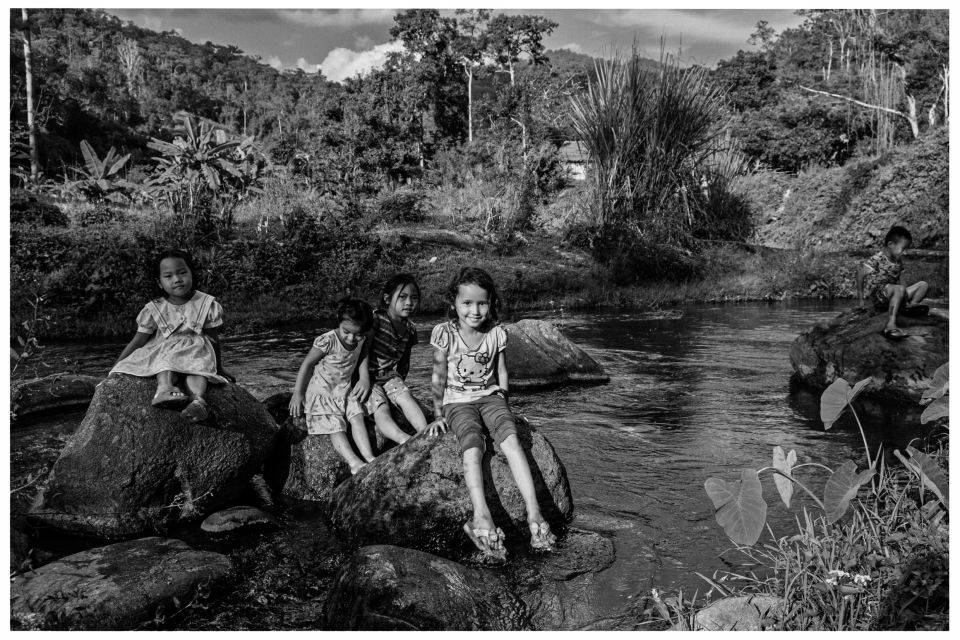 Kinder auf Felsen am Flussufer, umgeben von Vegetation und Bergen, in Schwarz-Weiß.