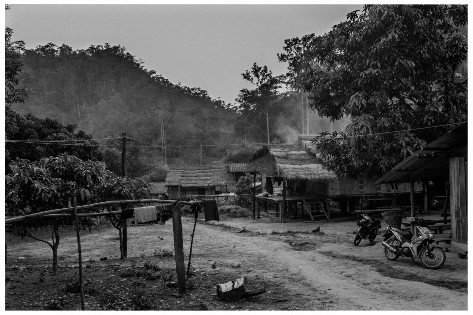 Ländliches Dorf mit Strohdächer, Motorrädern, Wäschestange, Hügel und Rauch im Hintergrund.