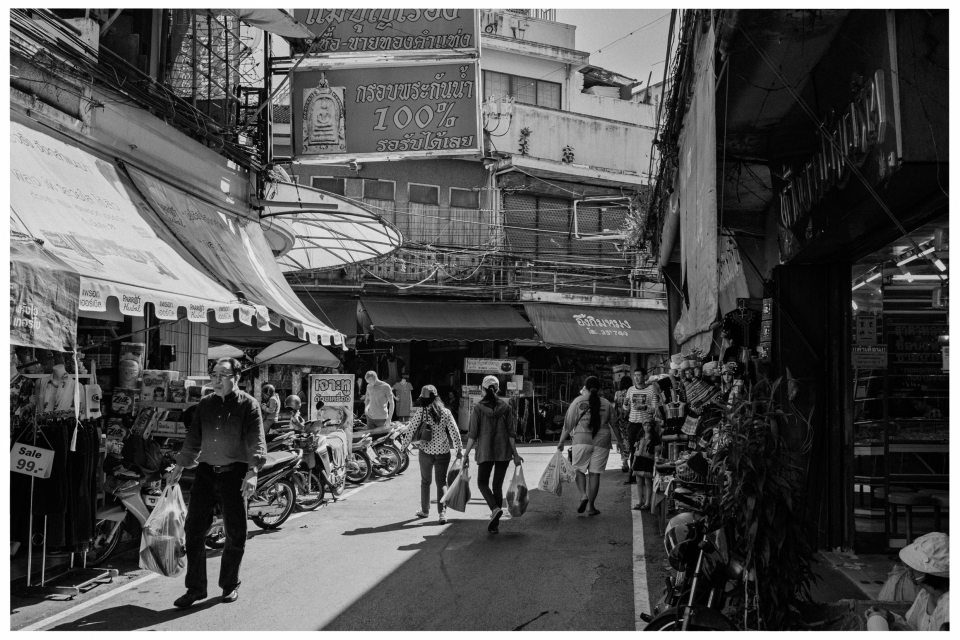 Geschäftiger Markt in Schwarzweiß mit Personen, die einkaufen, Motorräder und Schilder sichtbar.
