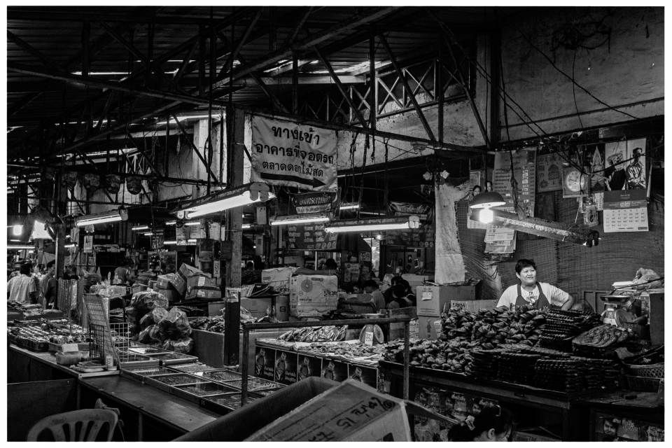 Marktstand in überdachtem Gebäude mit Lebensmitteln, Verkäufer und Schildern, gedämpftes Licht.